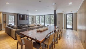 A spacious living room with a large wooden dining table, comfortable seating, a fireplace, and large windows offering a view of the outdoors. - Luxury Ski-In/Out Residence at Golden Peak (Vail)