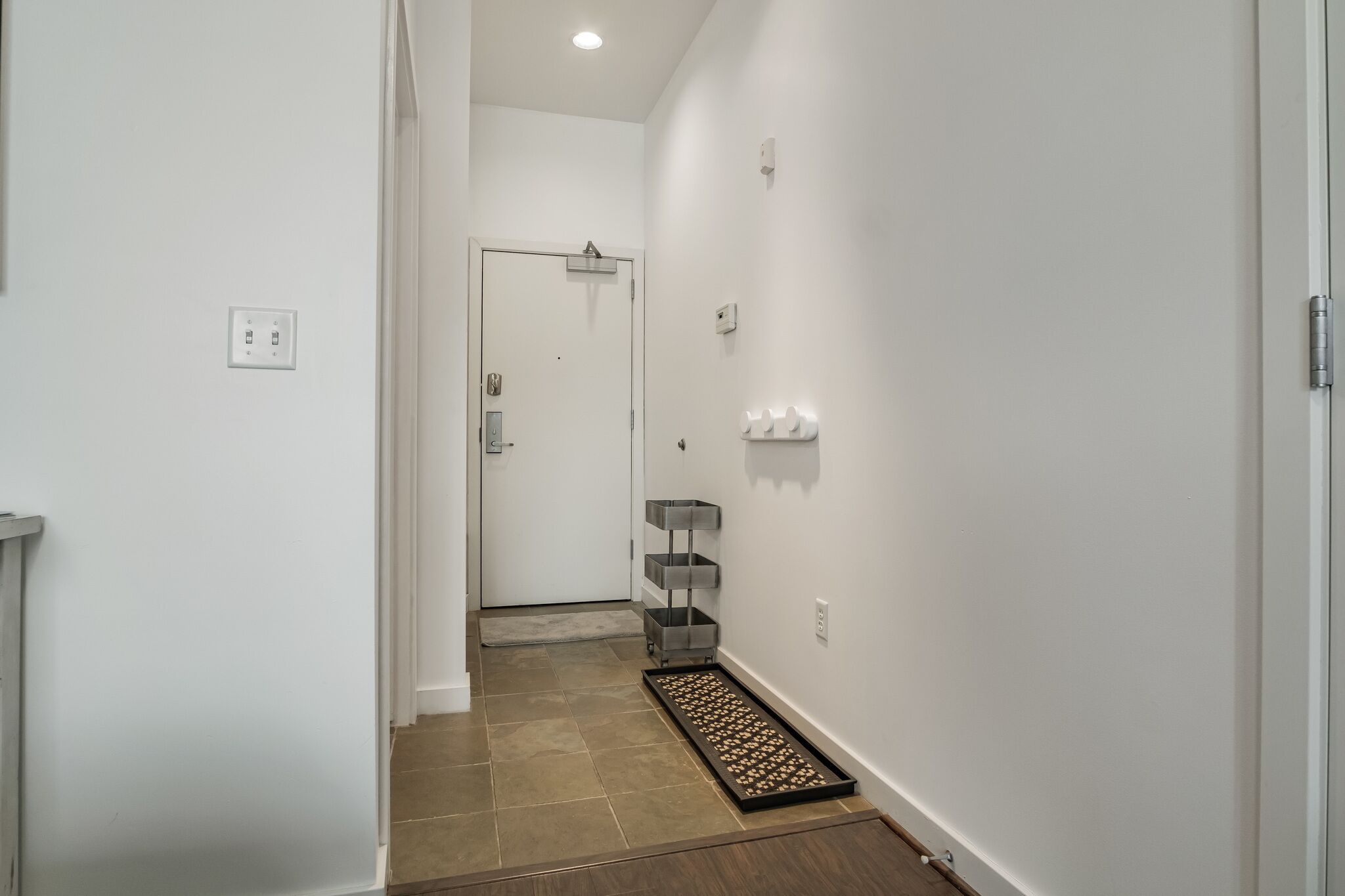 A narrow hallway with a door at the end, a small shelf unit, and a mat on the floor.