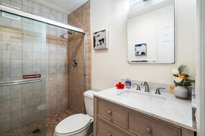 A bathroom with a glass shower enclosure, a toilet, a sink with a marble countertop, and a mirror above the sink. - Kona Isle D2 (Kailua-Kona)