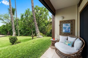 A wicker sofa with cushions and a small table with a decorative pillow, positioned on a balcony with a view of a well-maintained lawn and tall palm trees. - Kona Isle D2 (Kailua-Kona)