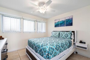 A bedroom with a bed, a ceiling fan, a nightstand, and a window with blinds. - Kona Isle D2 (Kailua-Kona)