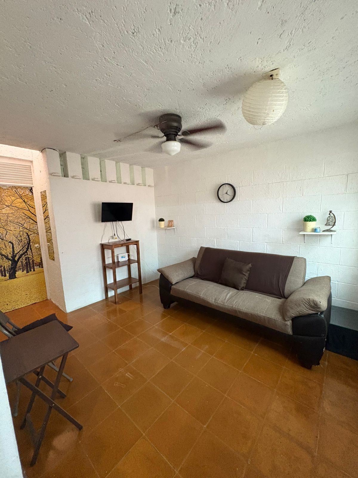 A living room with a sofa, a TV, a ceiling fan, a clock, and a small shelf with decorative items.