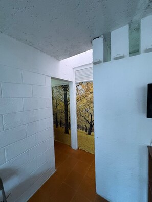 Un couloir aux murs en briques blanches, doté d’un sol carrelé et d’une porte donnant sur une pièce ornée d’une fresque représentant des arbres.