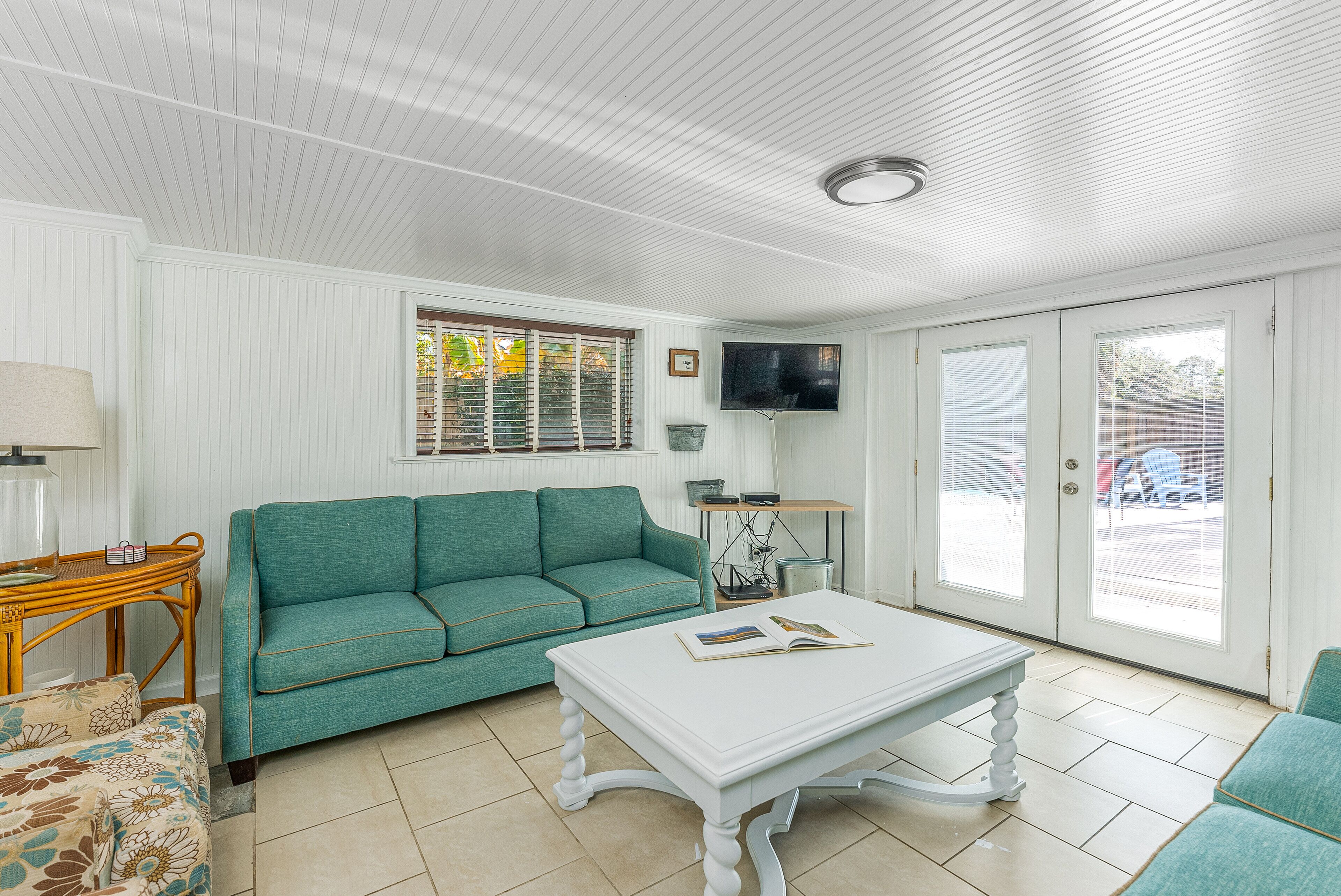 Un salon avec un canapé de couleur turquoise, une table basse blanche sur laquelle repose un livre, une table d’appoint en bois et un téléviseur fixé au mur.