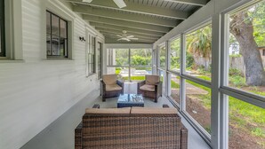 Eine überdachte Veranda mit Korbmöbeln, Deckenventilatoren und großen Fenstern, die einen Blick auf einen Garten bieten.