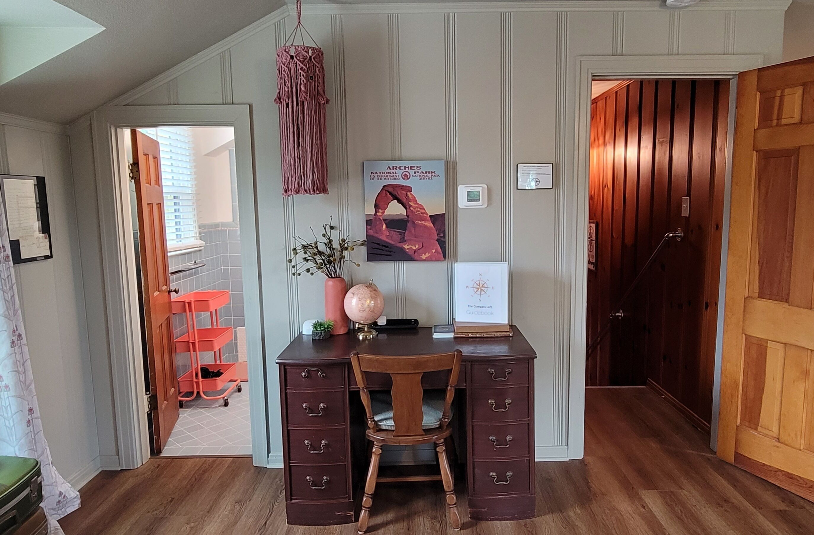 Un bureau en bois avec des tiroirs, une chaise, une décoration murale en macramé, un globe terrestre, une carte encadrée et une petite plante.