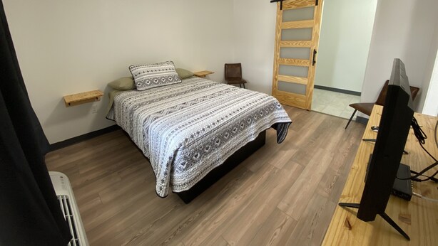 A bedroom with a bed, a desk, a chair, a heater, and a sliding door. - The Sage Motel (Saratoga)
