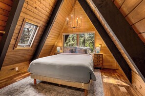 Ein Schlafzimmer mit schrägem Dach, einem Bett, einem Nachttisch, einer Lampe und einem Fenster mit Blick auf Bäume.