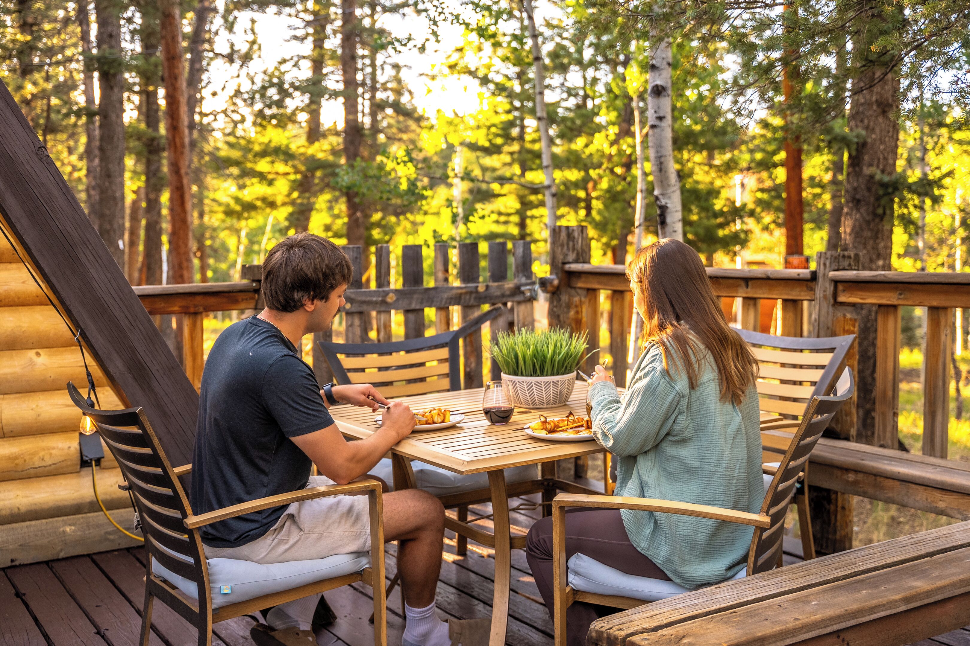A wooden deck with two people dining at a table, surrounded by wooden chairs and a small potted plant.