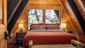 A bedroom with a sloped ceiling, a large bed with red bedding, two bedside lamps, and a view of trees through a window.