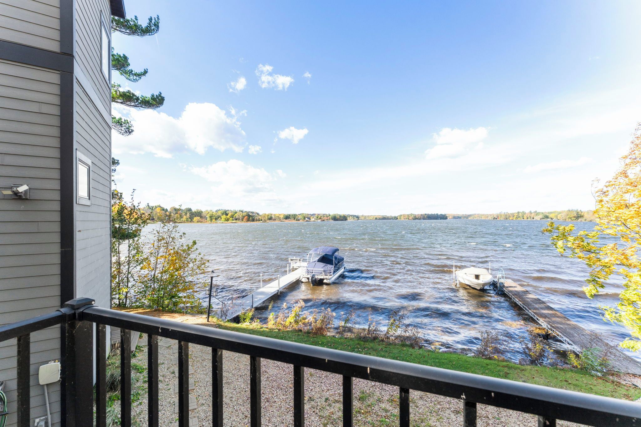 A lakeside view from a balcony with a dock extending into the water, a boat, and a house in the background.
