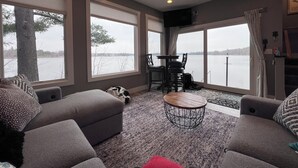 A living room with a large window overlooking a body of water, a ceiling fan, and a small round coffee table. - Lakefront Luxury Modern Home With Docks, Fire Pit & Pontoon Boat Rental (Turtle Lake)