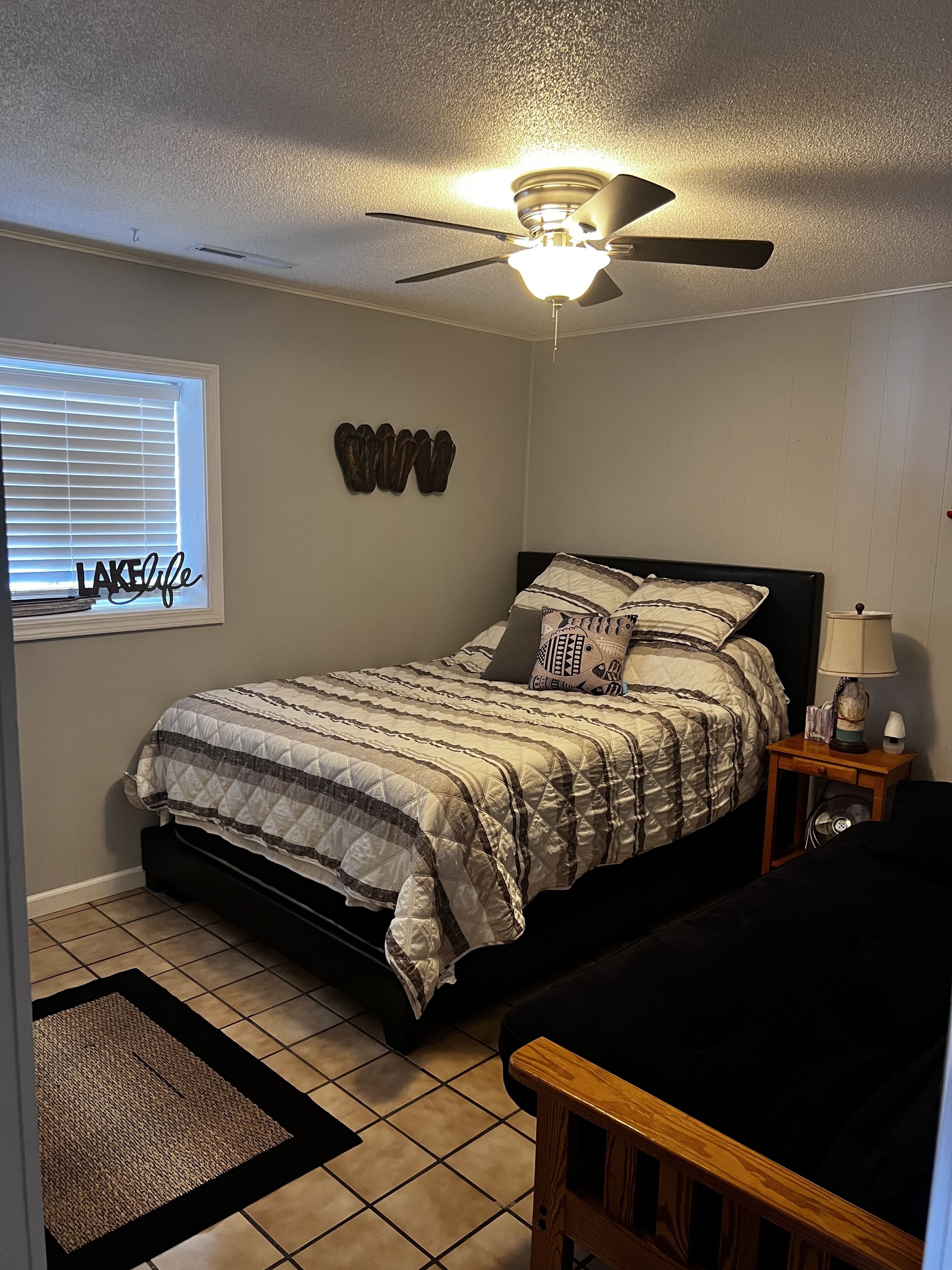 A bedroom with a bed, a ceiling fan, a bedside table with a lamp, a window with blinds, and a rug.