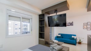 A modern hotel room with a bed, a blue sofa, a flat-screen TV mounted on the wall, a window with blinds, and a small table with towels. - Bright Studio - GRU (Guarulhos)