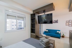 Een moderne hotelkamer met een bed, een blauwe bank, een flatscreen televisie aan de muur, een raam met zonwering en een klein tafeltje met handdoeken.