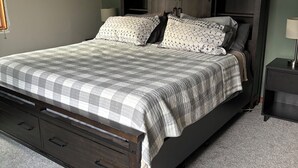 A bedroom with a dark wooden bed, a checkered bedspread, and a nightstand with a lamp. - Spacious Home that is close to EAA and Shopping. (Oshkosh)
