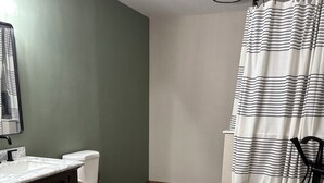 A bathroom with a wooden floor, a white toilet, a dark wooden vanity, a white sink, and a shower with a striped curtain. - Spacious Home that is close to EAA and Shopping. (Oshkosh)