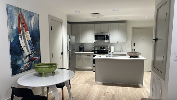A modern kitchen with white cabinets, a central island, a dining area with a table and chairs, and a painting of a sailboat on the wall. - Easy walk to restaurants, coffee and ice cream shops, retail shopping, and more! (Tupelo)