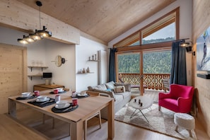 A cozy room with a wooden dining table set for four, a sofa, a red chair, and a large window with a view of a forested mountain. - PERLE DE SAVOIE A402 (CHATEL)