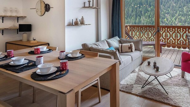 A dining area with a table set for four, a sofa, a red chair, and a balcony with a view of a forest. - PERLE DE SAVOIE A402 (CHATEL)