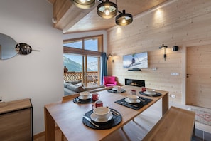 A wooden interior with a dining table set for four, a television mounted on the wall, and a view of the mountains through large windows.