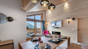 A cozy room with a wooden dining table set for four, a TV mounted on the wall, and a view of the mountains through large windows.