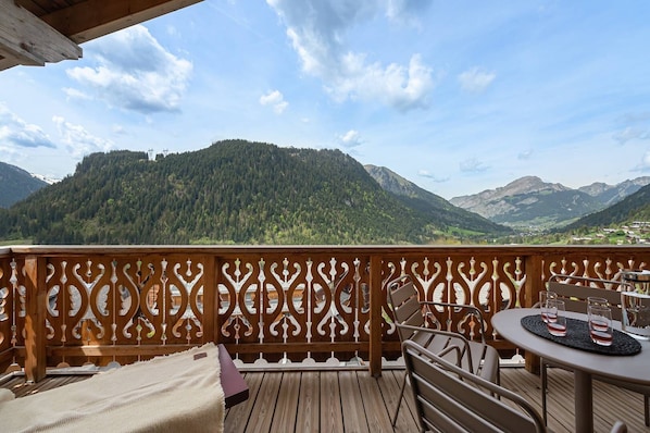 A wooden deck with a view of mountains, a table with glasses, and a chair.