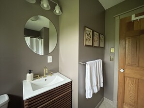 Ein Badezimmer mit einem weißen Waschbecken, einem runden Spiegel und einer Holzausstattung. An der Wand hängen drei gerahmte Bilder und ein Handtuchhalter mit Handtüchern.