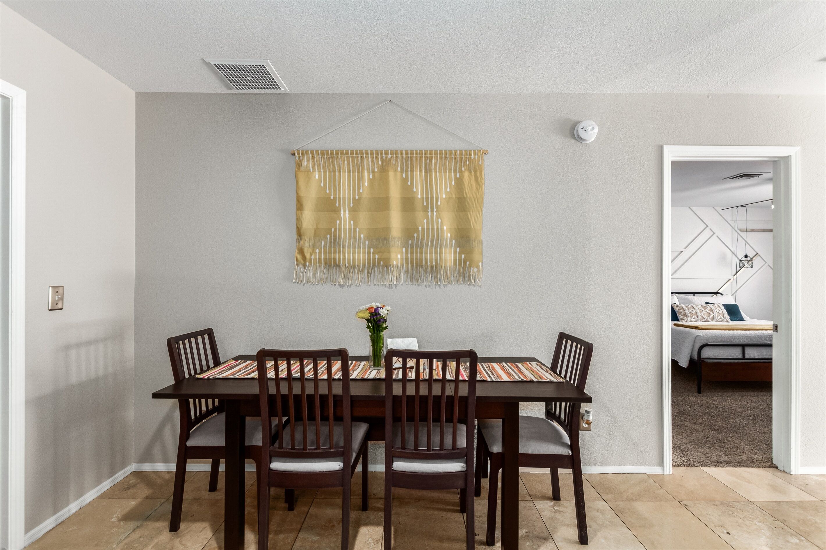 A dining area with a table set for four, a hanging decorative piece, and a view into a bedroom with a bed and a nightstand.