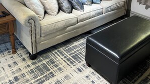 Un salon avec un canapé beige, de nombreux coussins, un pouf noir et des œuvres d’art murales représentant des animaux.
