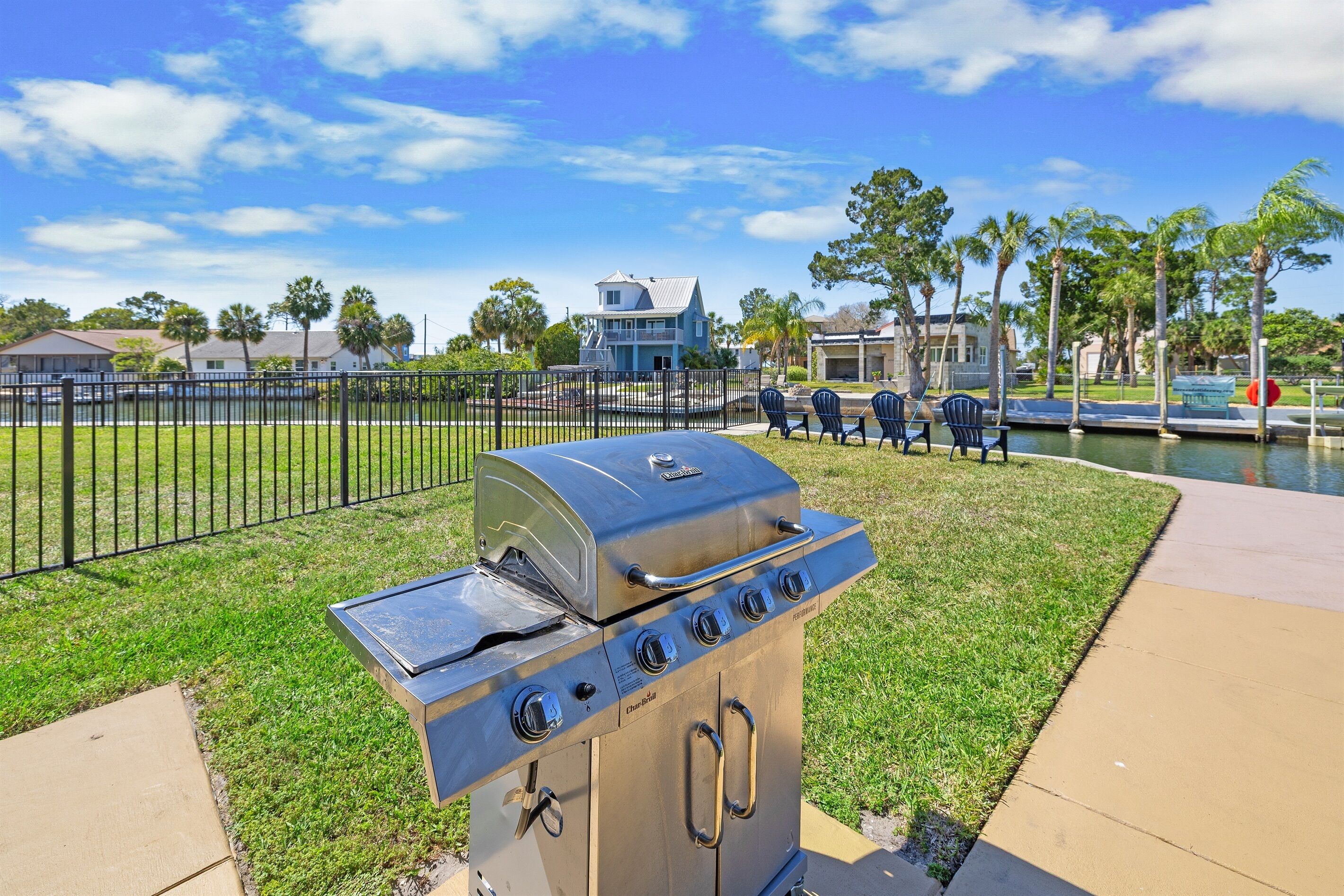 A stainless steel barbecue grill with a chimney, located on a grassy area with a fence and a body of water in the background.