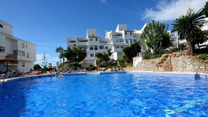 Una piscina con agua cristalina azul, rodeada de sillones de descanso y sombrillas, con edificios al fondo.