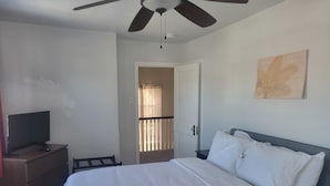 Un dormitorio con una cama, un televisor sobre un aparador de madera, un ventilador de techo y una puerta que da a un balcón.
