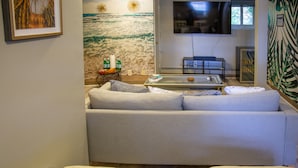 Una sala de estar con un sofá, un reposapiés y una mesa de centro de vidrio. Hay un televisor montado en la pared y un mural decorativo.