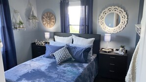 Une chambre à coucher comprenant un lit, deux tables de chevet, une horloge murale, un miroir et une fenêtre avec des rideaux.