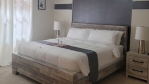 Ein Schlafzimmer mit hölzernem Bettgestell, Deckenventilator, zwei Nachttischen mit Lampen, Wandbild und einem Fenster mit Vorhängen.
