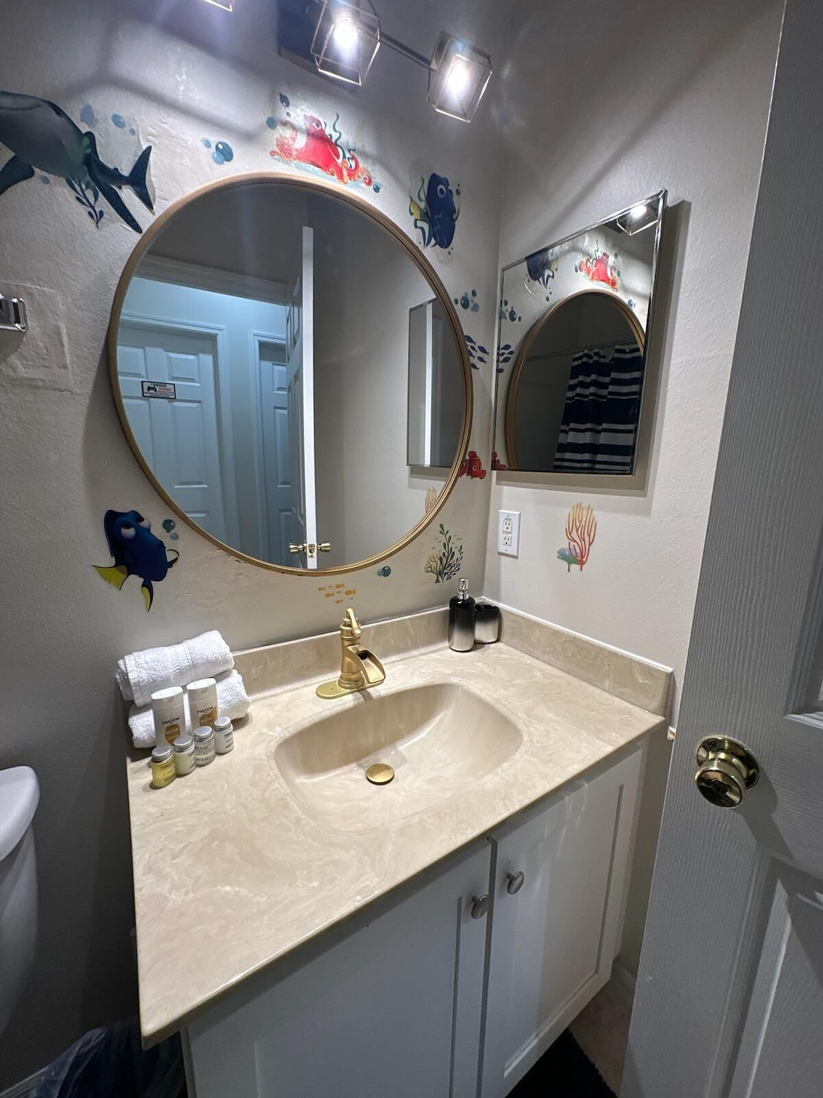 A bathroom with a beige vanity, a gold faucet, and a large mirror. There are decorative fish decals on the wall and a small shelf with toiletries.
