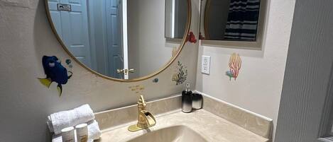 A bathroom with a beige vanity, a gold faucet, and a large mirror. There are decorative fish decals on the wall and a small shelf with toiletries.