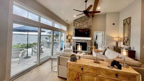 A spacious living room with a stone fireplace, a ceiling fan, and a large TV. - Brand new private  beachfront home in Mashpee on Waquoit Bay (Mashpee)