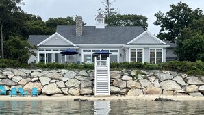 A waterfront property with a stone wall, a staircase leading to a deck, and a row of blue chairs. - Brand new private  beachfront home in Mashpee on Waquoit Bay (Mashpee)