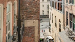 Une ruelle étroite, bordée de bâtiments, avec un mur de briques d’un côté et un immeuble moderne aux grandes fenêtres de l’autre.