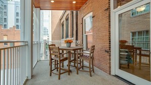 Een balkon met een houten tafel en stoelen, een bloempot en uitzicht op omliggende gebouwen.
