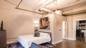 Quarto moderno com uma cama, uma escrivaninha e uma cadeira. Há um quadro na parede e um abajur ao lado da cama.