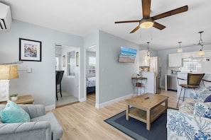 A modern living room with a ceiling fan, a dining area, a kitchen, and a bedroom with a large window. - Flagler Ave 1 Bedroom/1 Bath -Eat, Drink, Play! (New Smyrna Beach)