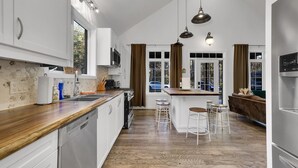 Una cocina moderna con armarios blancos, encimera de madera y una isla central con taburetes. Hay luces colgantes sobre la isla y un ventanal con cortinas.
