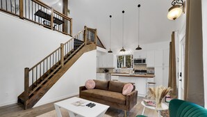 Un salón espacioso con una escalera de madera, un sofá verde, una mesa de centro blanca y una cocina al fondo.