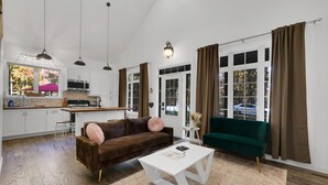 Una sala moderna con un sofá marrón, una mesa de centro blanca y un sillón verde. Se ve una cocina con alacenas blancas y un microondas al fondo.