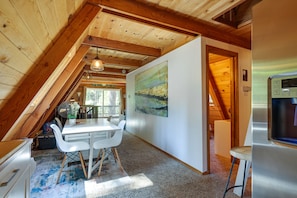 A cozy attic bedroom with a wooden ceiling, a dining table with chairs, a painting on the wall, and a refrigerator.