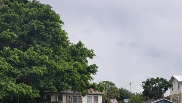 Une jetée en bois mène à une maison avec un petit abri et deux chaises vertes, près de l’eau.