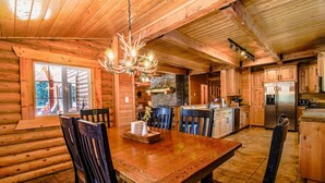 Interior de una cabaña de troncos con una mesa de comedor de madera, sillas, un candelabro y una zona de cocina.